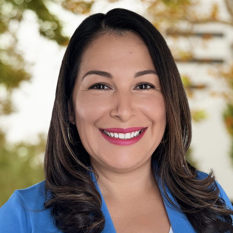 Portrait of a smiling woman with long dark hair wearing a black top, blurred background with artwork on the wall.