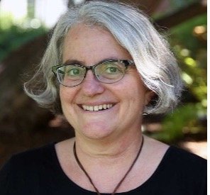 A smiling person with glasses and short gray hair wearing a black shirt, standing outdoors with blurred greenery in the background.