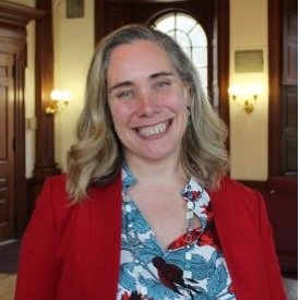 A smiling person with shoulder-length blond hair, wearing a red blazer and a floral blouse, standing in a room with wood paneling and arched windows.