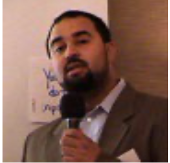 A man speaking into a microphone at a conference or meeting, wearing a suit jacket and standing in front of a wall with writing on it.